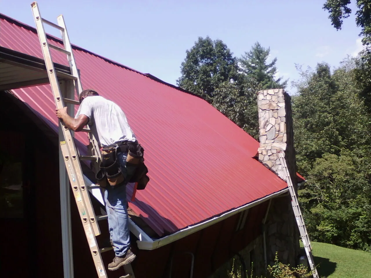 Roofer on a ladder beside a red metal roof and stone chimney — Soffit & Fascia Repair by Roofing Brothers in Coin